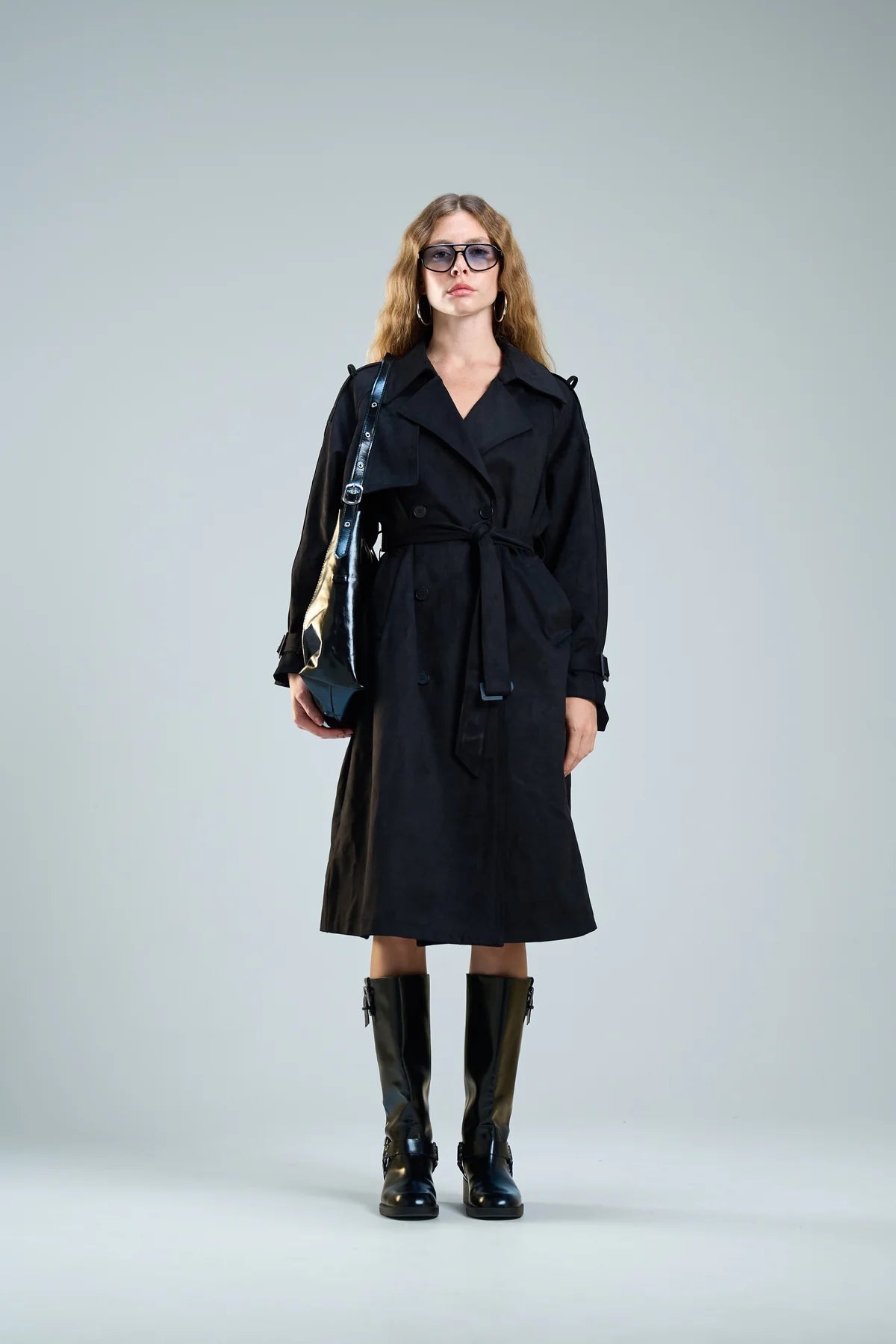 אישה עם מעיל טרנץ' שחור ארוך, מגפיים גבוהים ותיק צד שחור, לוק חורפי אלגנטי.