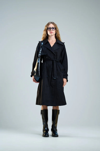 אישה עם מעיל טרנץ' שחור ארוך, מגפיים גבוהים ותיק צד שחור, לוק חורפי אלגנטי.